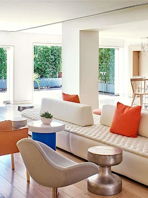 A bright, modern living area with white sectional sofa, colorful cushions, stylish chairs, round tables, and large windows letting in natural light.
